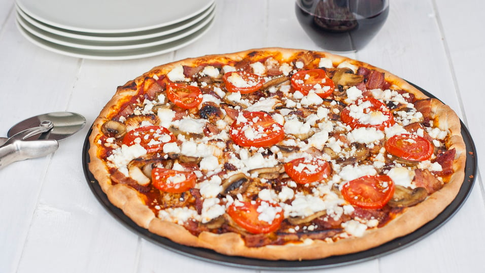 Wide shot of cooked pizza topped with BBQ chicken, bacon, feta and tomatoes.