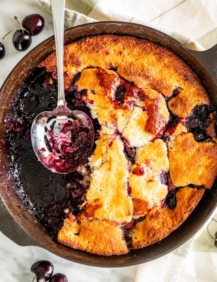 overhead shot of cherry cobbler in a pan with pieces missing. A serving spoon is resting in the empty pan space
