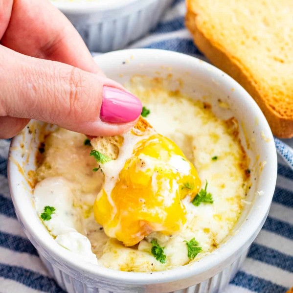 a hand dunking a piece of toast in a ramekin with creamy baked eggs