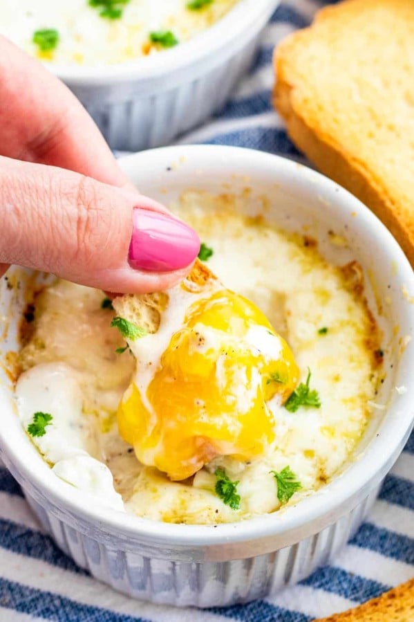 a hand dunking a piece of toast in a ramekin with creamy baked eggs
