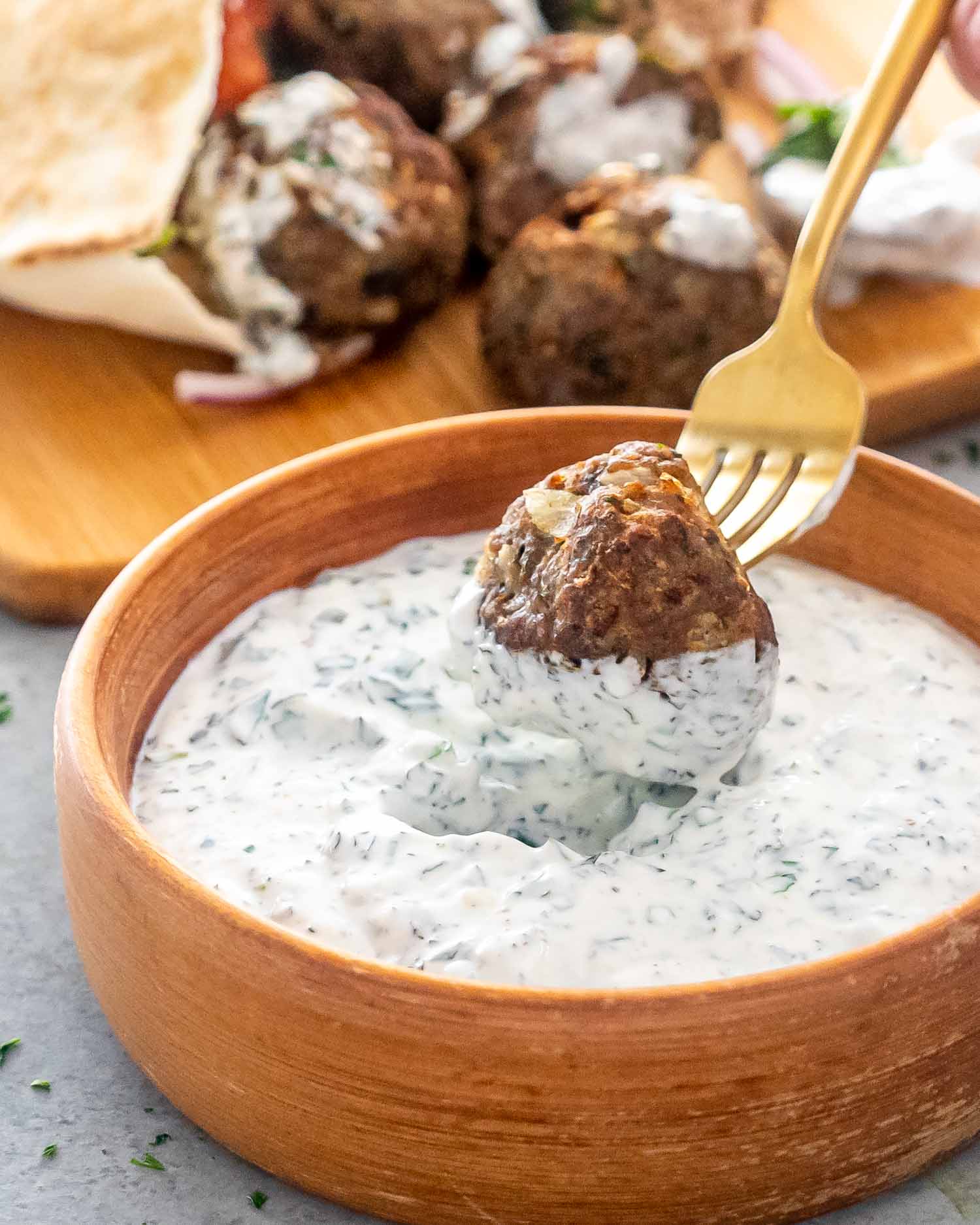 a fork dipping a lamb meatball in a bowl with yogurt mint sauce.