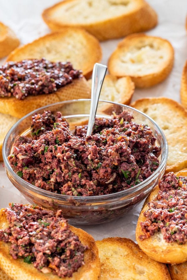 side view shot of a bowl of olive tapenade with a serving utensil in it, surrounded by pieces of toasted baguette