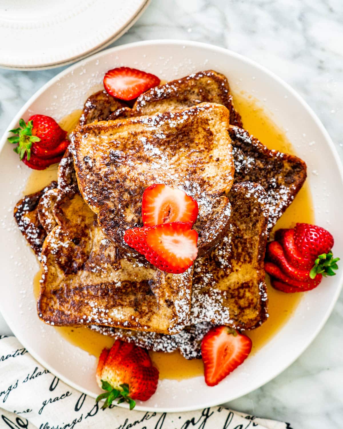 overhead shot of french toast on a white plate garnished with strawberries and drizzled with maple syrup