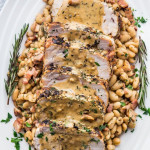 overhead shot of sliced rosemary garlic pork roast covered in gravy on top of white beans on a serving plate garnished with rosemary sprigs