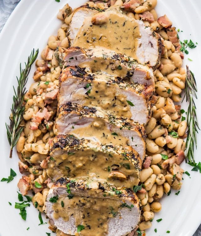 overhead shot of sliced rosemary garlic pork roast covered in gravy on top of white beans on a serving plate garnished with rosemary sprigs