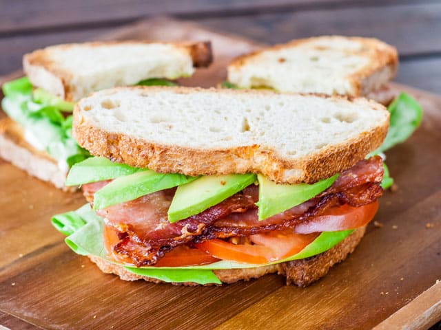 one avocado blt sandwich on a cutting board with one avocado blt cut in half in the background