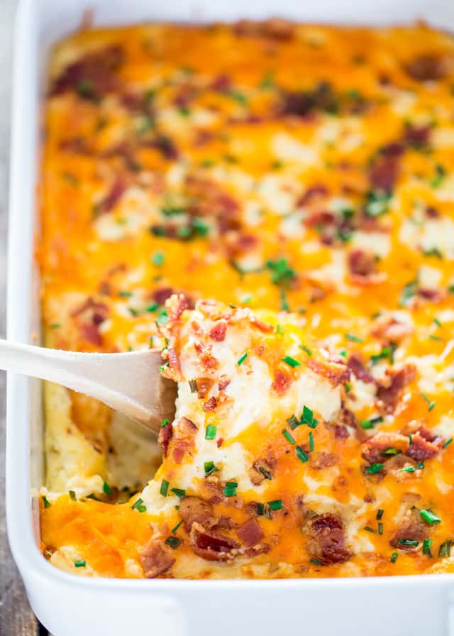 cheesy mashed potatoes with bacon and chives in a casserole dish with a wooden spoon