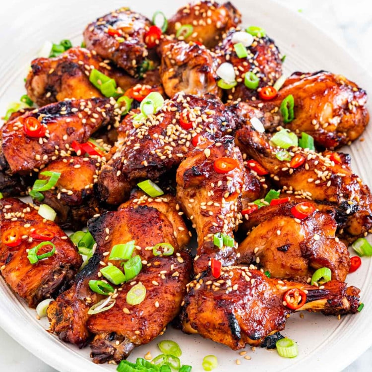 side view shot of a plate of sticky Chinese chicken wings, topped with sesame seeds, chopped green onion and sliced red chilis