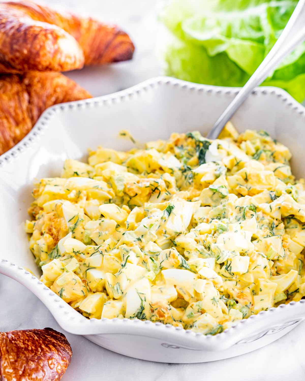 egg salad in a white bowl with croissants around it and lettuce