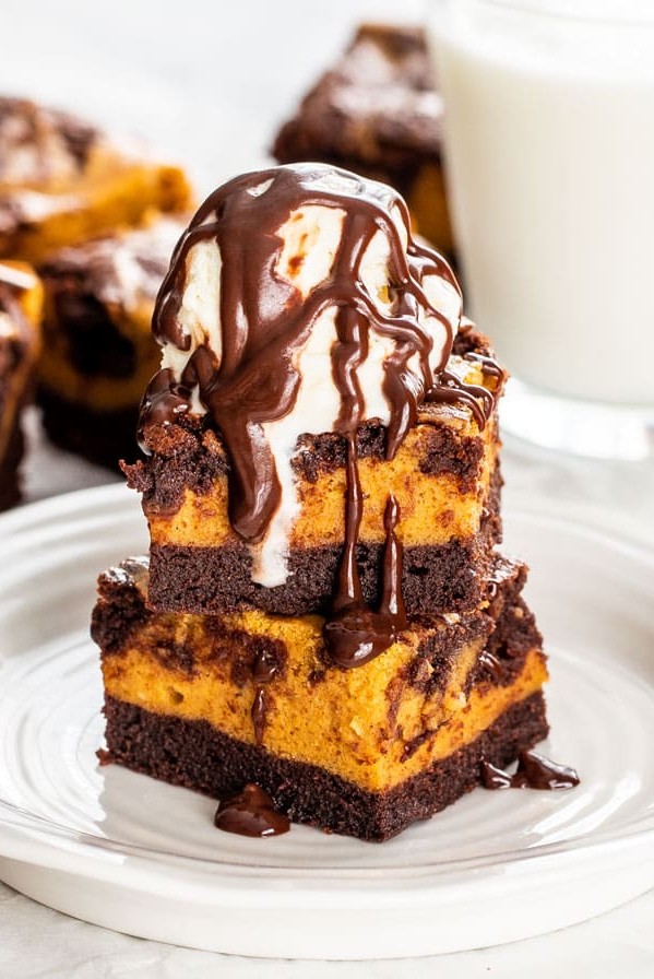 pumpkin cheesecake brownies stacked on a plate. Brownies are topped with a scoop of vanilla ice cream and drizzled in chocolate sauce