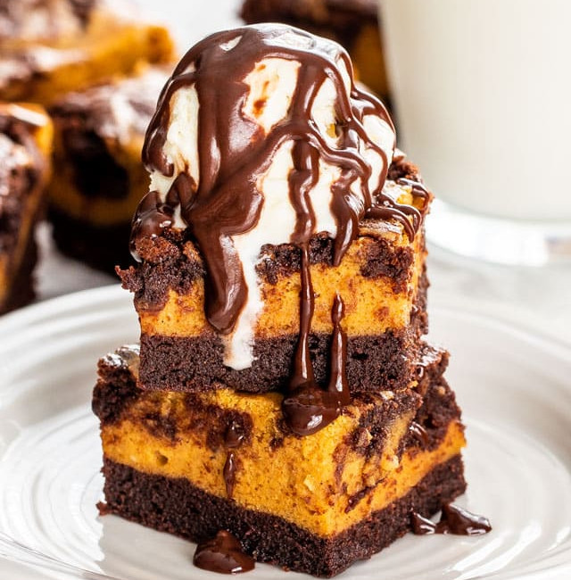 pumpkin cheesecake brownies stacked on a plate. Brownies are topped with a scoop of vanilla ice cream and drizzled in chocolate sauce