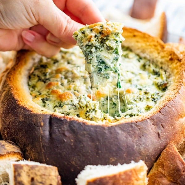 a hand dipping a piece of bread in a spinach and artichoke dip in a bread bowl.