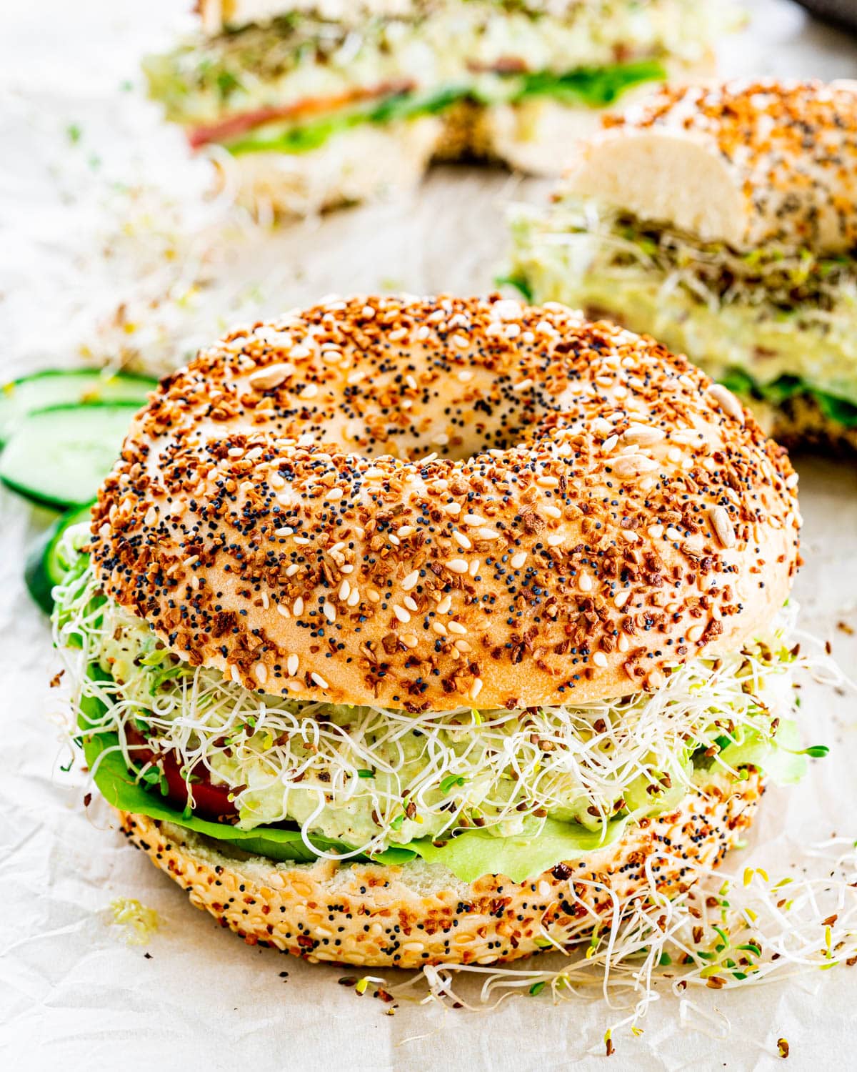 a bagel loaded with avocado egg salad, lettuce, tomatoes, cucumber and alfalfa sprouts