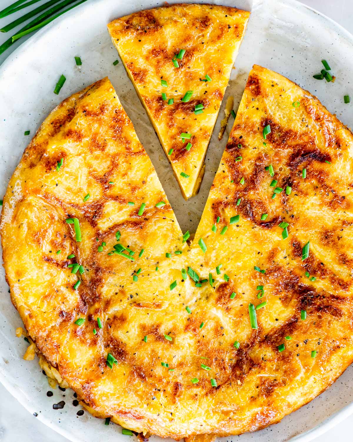 overhead shot of spanish omelette on a plate with one slice cut out, and garnished with chives