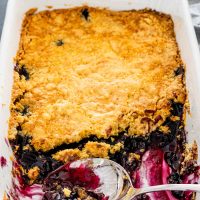 blueberry dump cake in a white baking dish with almost half of it out and a serving spoon in it.