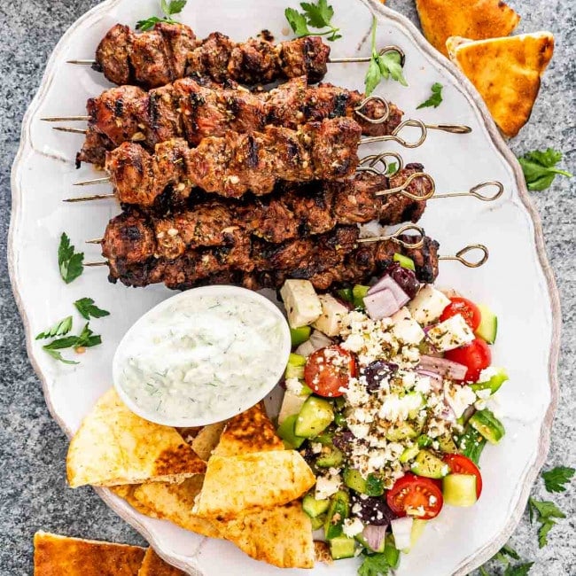 lamb souvlaki on a platter with tzatziki sauce, greek salad and pita bread.