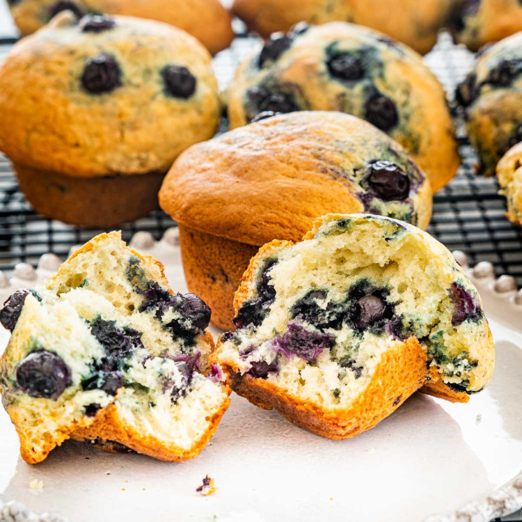 2 blueberry muffins on a plate, 1 cut in half with center exposed. More blueberry muffins in the background