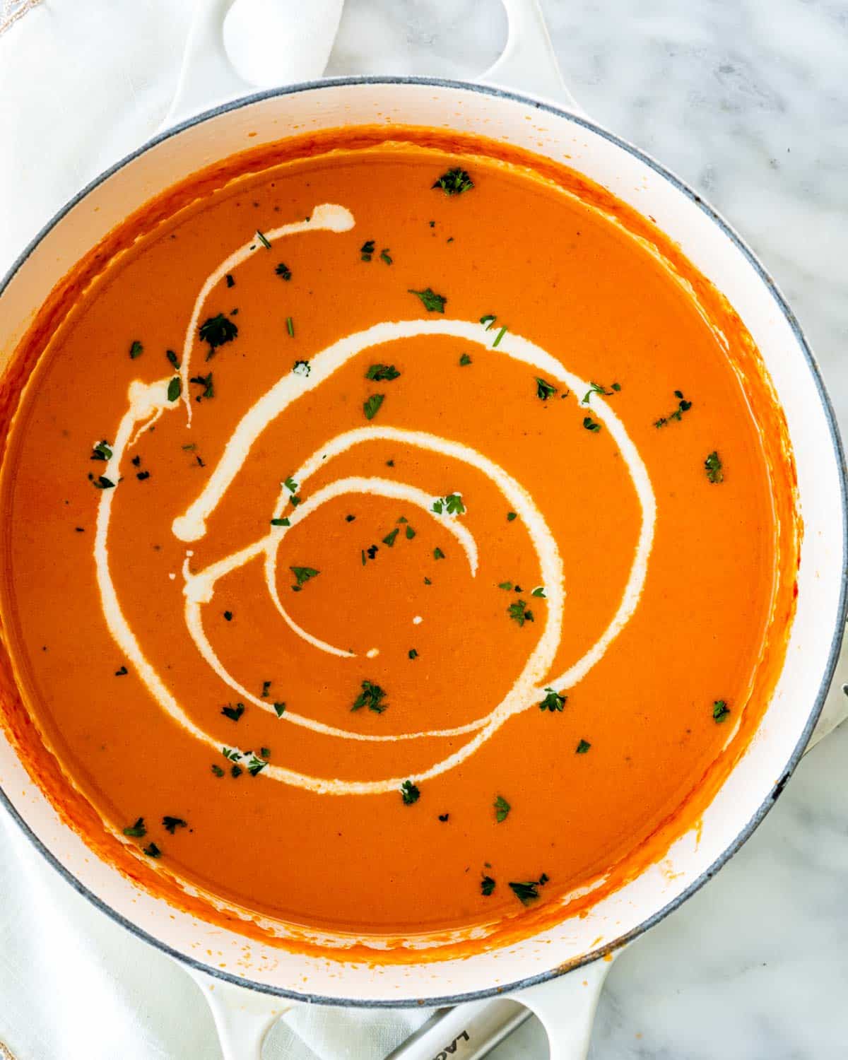 overhead shot of tomato bisque soup in a beige dutch oven with a drizzle of heavy cream garnished with parsley