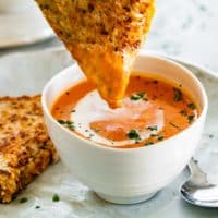 side view shot of tomato bisque in a soup bowl with a a hand dipping half a grilled cheese in the soup