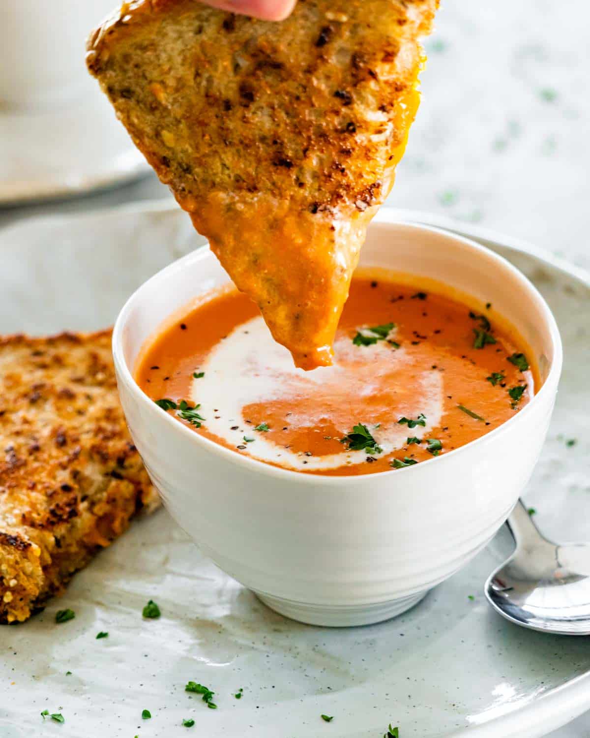 side view shot of tomato bisque in a soup bowl with a a hand dipping half a grilled cheese in the soup