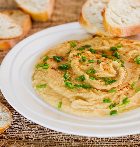 a bowl of roasted garlic hummus surrounded by slices of bread slices