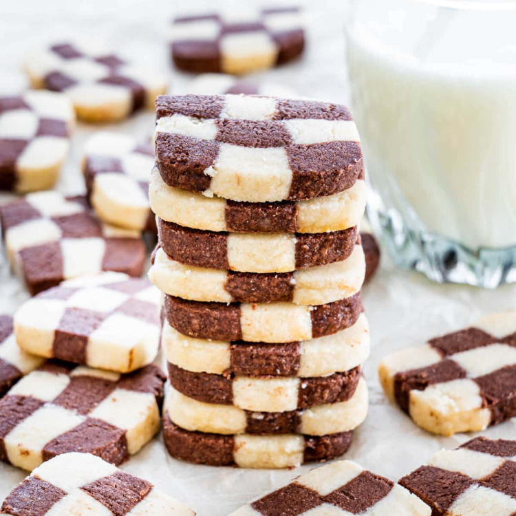 checkerboard cookies stacked on top of one another, surrounded by more cookies and a glass of milk in the background
