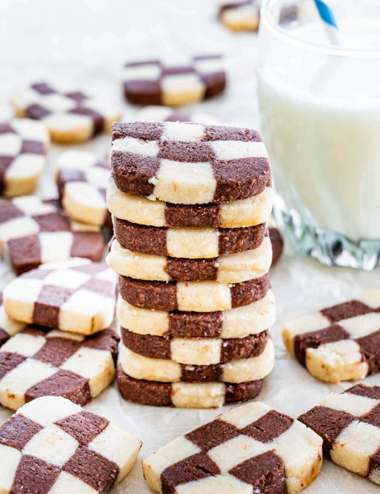 checkerboard cookies stacked on top of one another, surrounded by more cookies and a glass of milk in the background