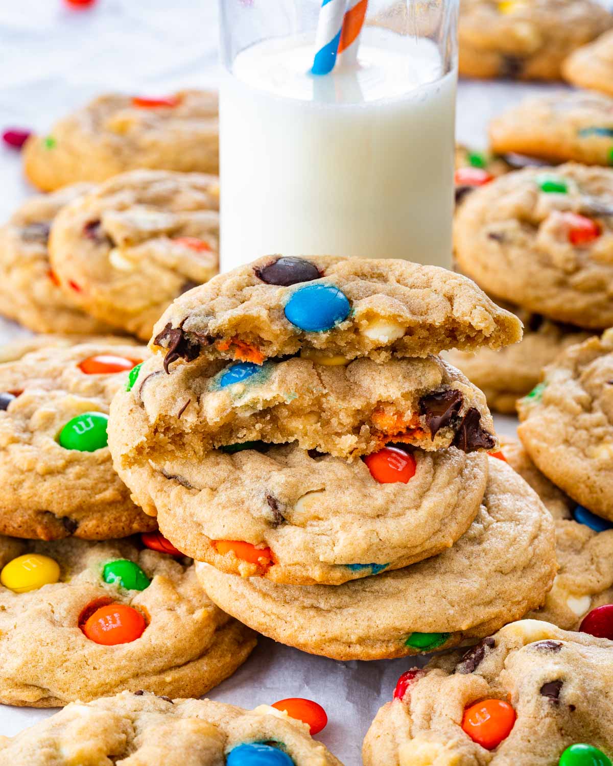 a bunch of m&m cookies and a glass of milk with the top cookie broken in half.