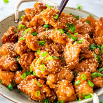 a pair of chopsticks picking of up piece of chicken from a bowl full of sesame chicken