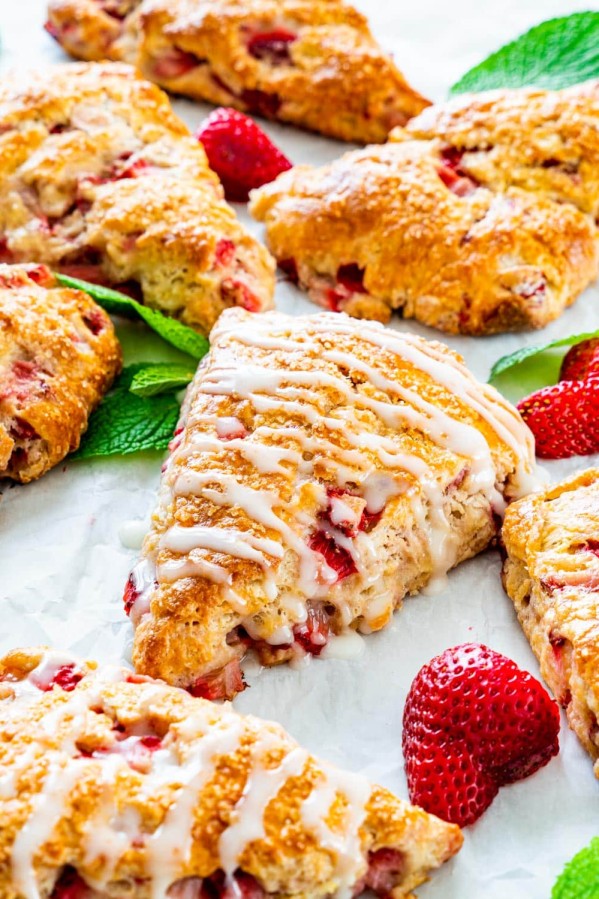 a bunch of strawberry scones drizzled with lemon icing on a piece of parchment paper, with scattered strawberries and mint