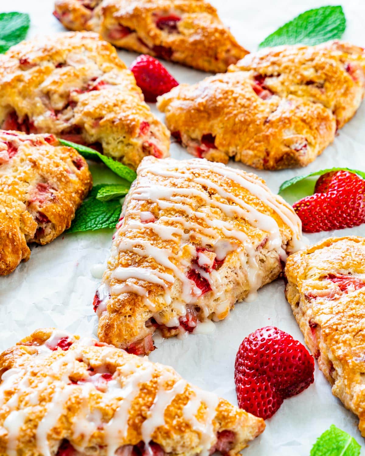 a bunch of strawberry scones drizzled with lemon icing on a piece of parchment paper, with scattered strawberries and mint