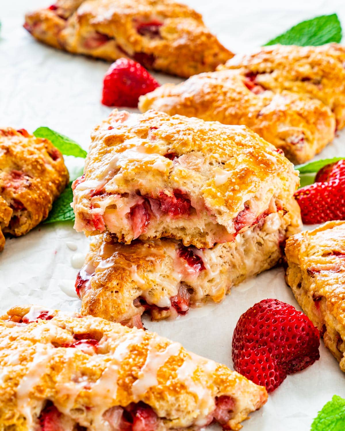 a bunch of strawberry scones drizzled with lemon icing on a piece of parchment paper with one scone broken in half, with each half stacked on top of each other