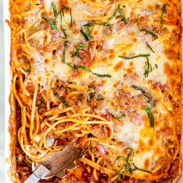 overhead shot of baked spaghetti casserole in a baking dish with a portion missing and a serving spoon resting in its place