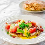 a plate of corn and chive pancakes with bacon and eggs topped with avocado and tomatoes