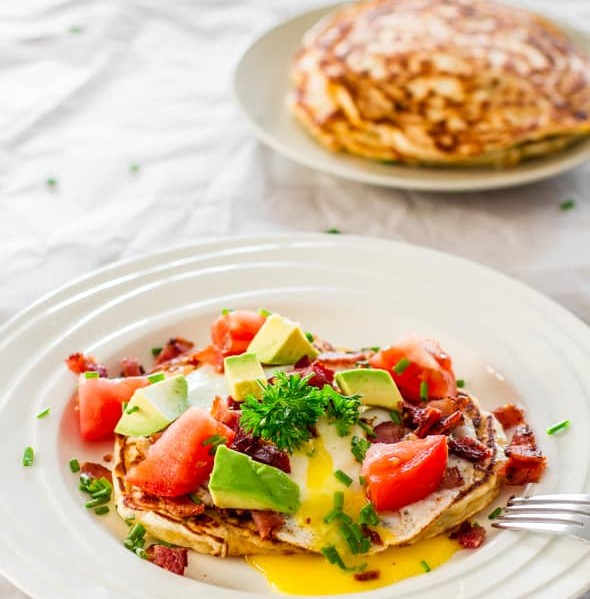 a plate of corn and chive pancakes with bacon and eggs topped with avocado and tomatoes