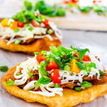 two navajo tacos on a piece of parchment paper with a cutting board with ingredients in the background