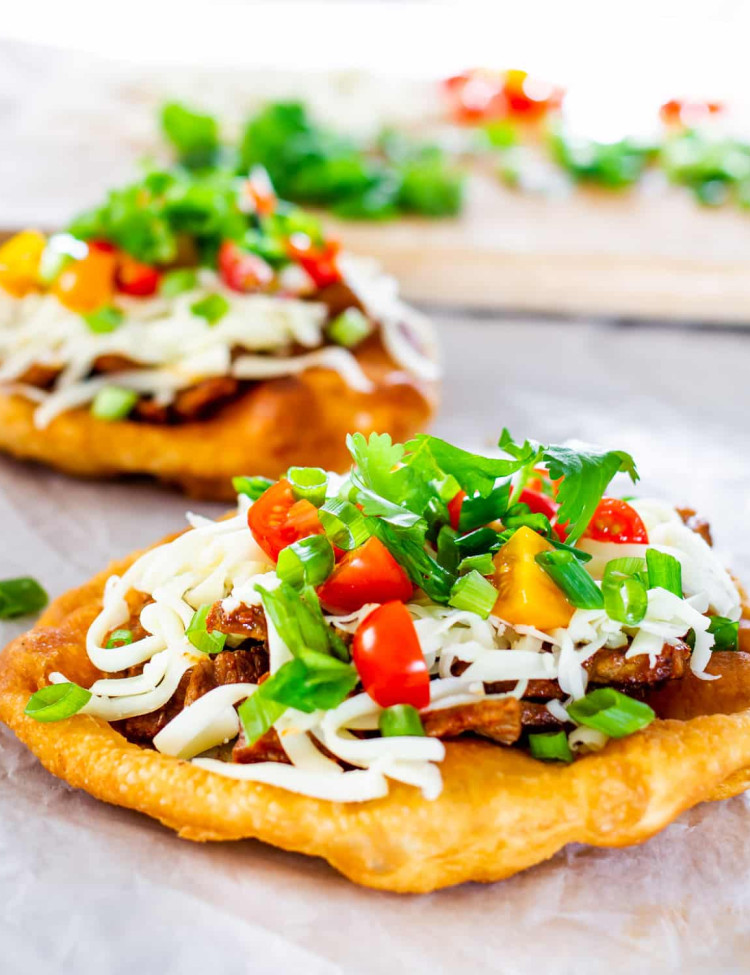 two navajo tacos on a piece of parchment paper with a cutting board with ingredients in the background