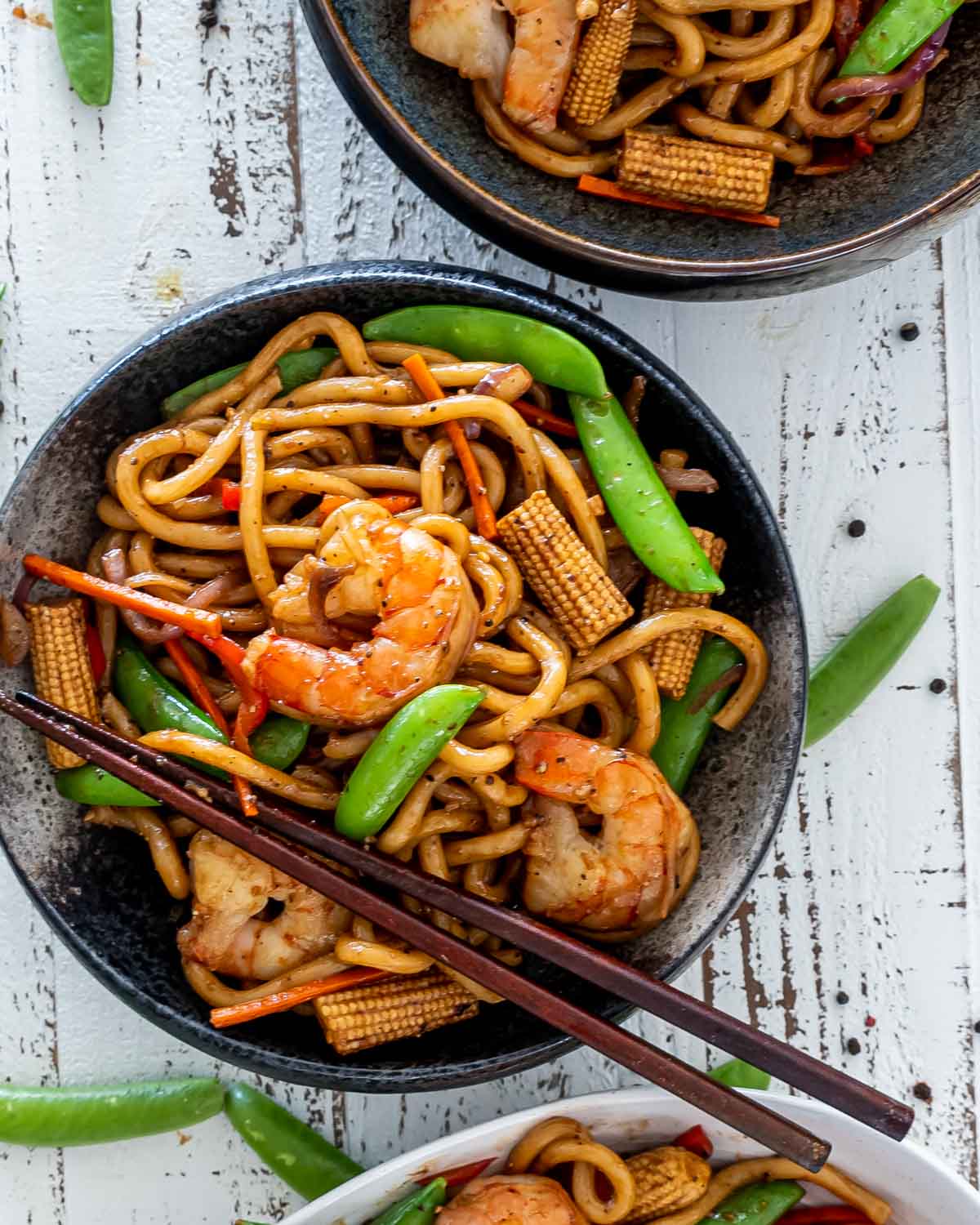 black pepper udon noodles with shrimp on a black plate with a pair of chopsticks.
