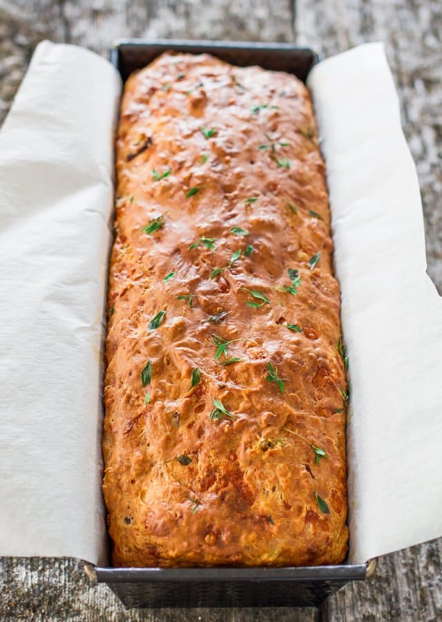 A loaf of Cheddar Olive Thyme Bread in a baking pan lined with parchment paper