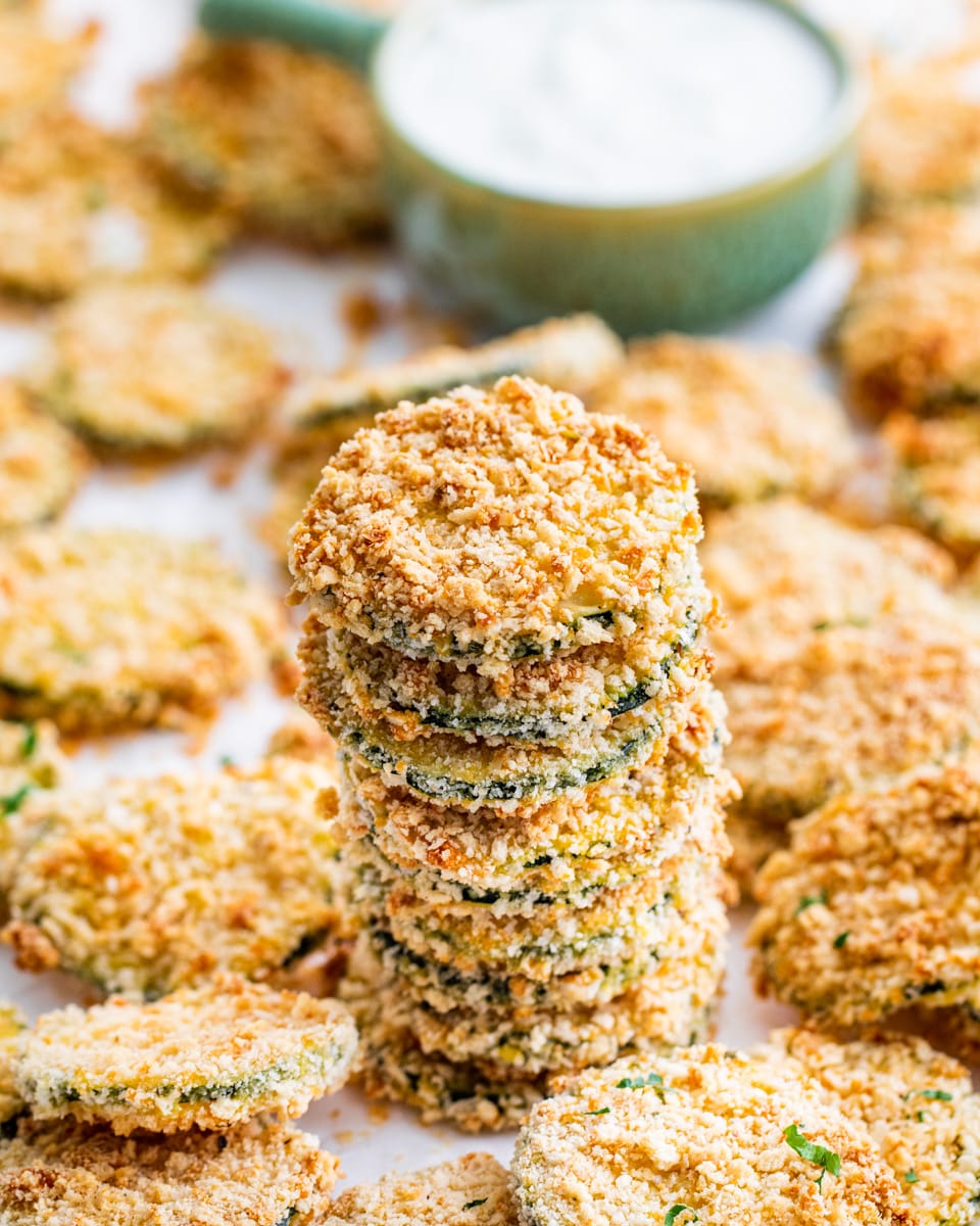 a stack of freshly baked zucchini crisps surrounded by lots of other crisps with a bowl of ranch dressing in the background