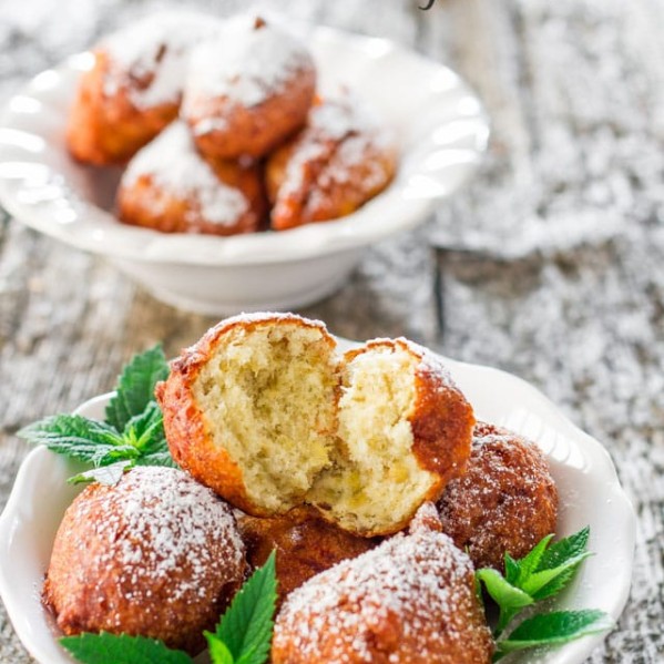 a bowl of banana fritters dusted in powdered sugar with one ripped in half