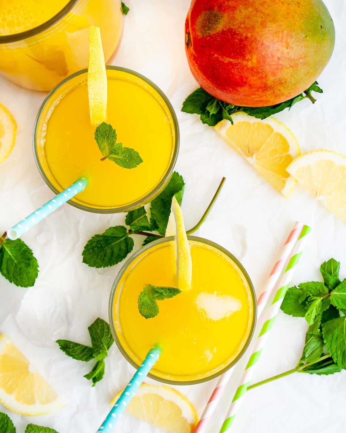 two glasses filled with mango lemonade with a mango next to them garnished with lemon slices and a mint leaf