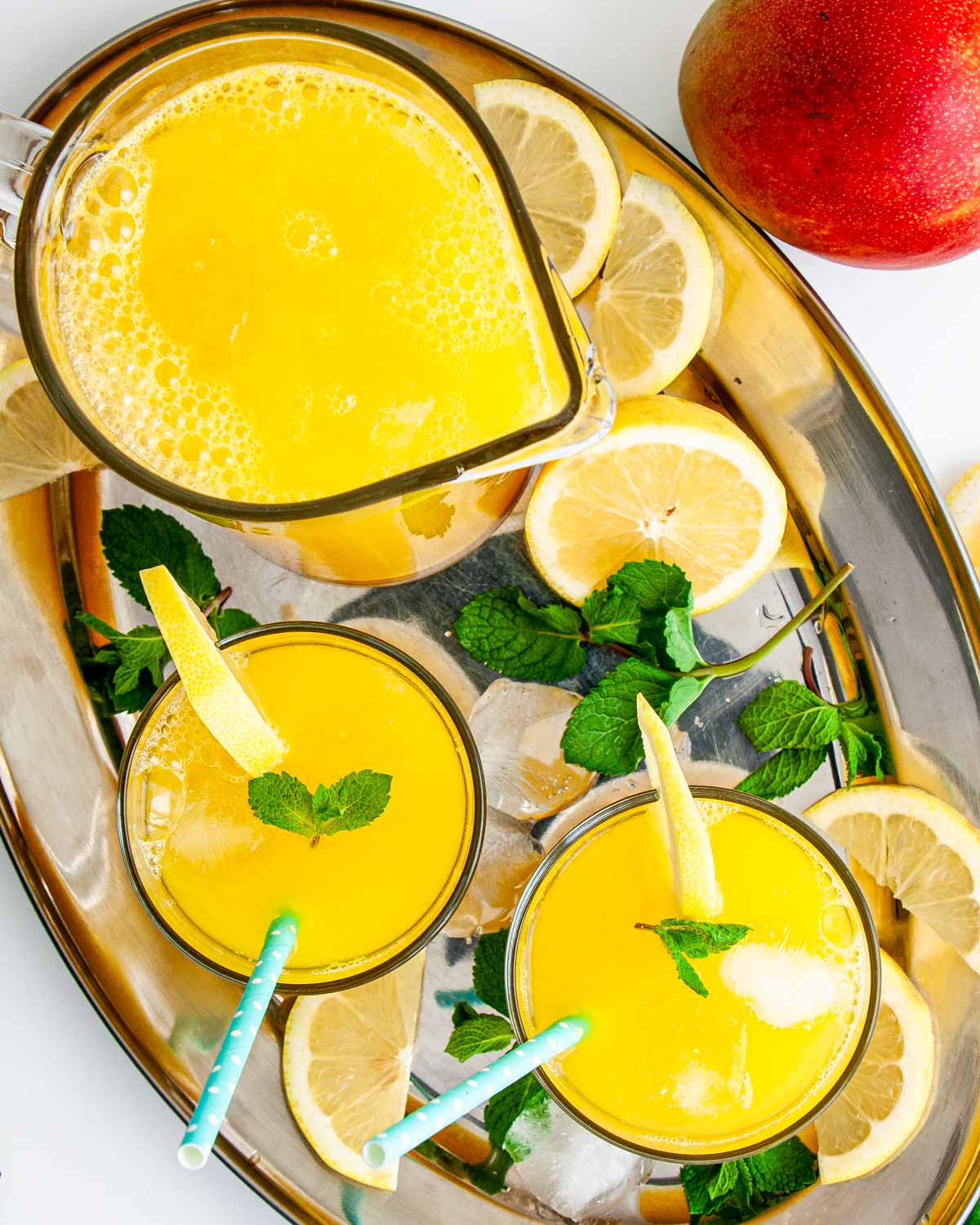a pitcher and two glasses filled with mango lemonade on a serving tray garnished with lemon slices and mint