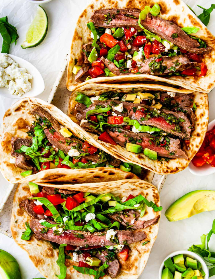 overhead shot of steak tacos on a platter with chimichurri sauce and garnished with tomatoes, avocado and feta