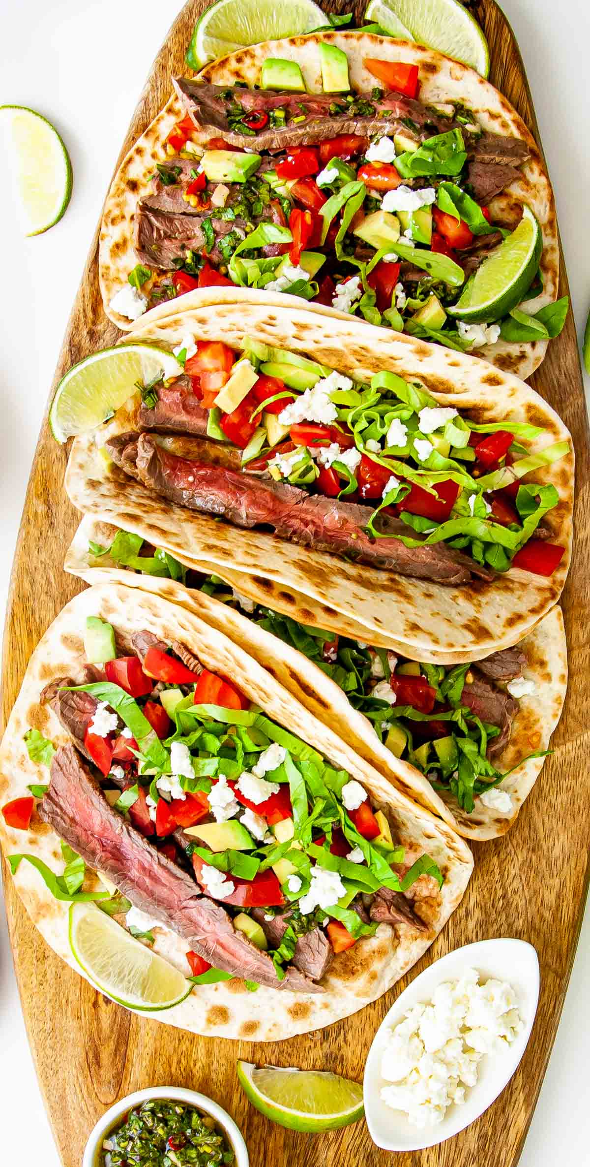 overhead shot of steak tacos on a cutting board with chimichurri sauce and garnished with tomatoes, avocado and feta