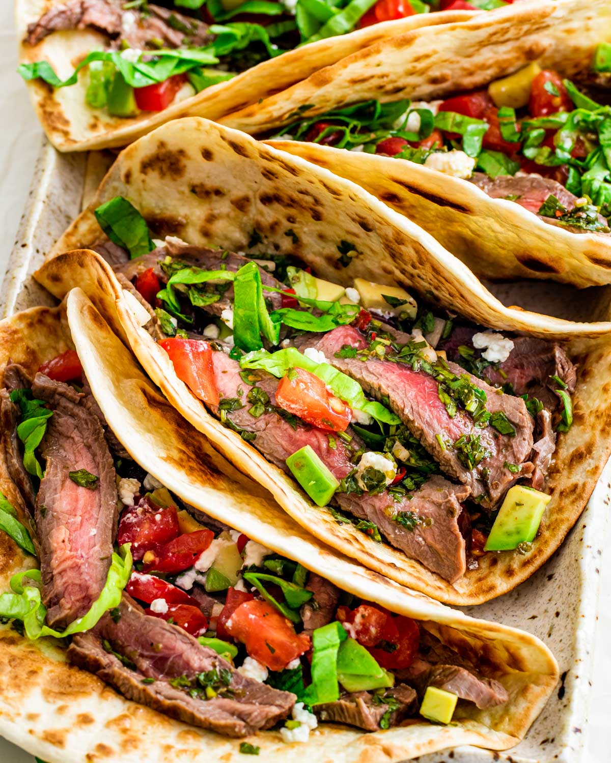 overhead shot of steak tacos on a platter with chimichurri sauce and garnished with tomatoes, avocado and feta