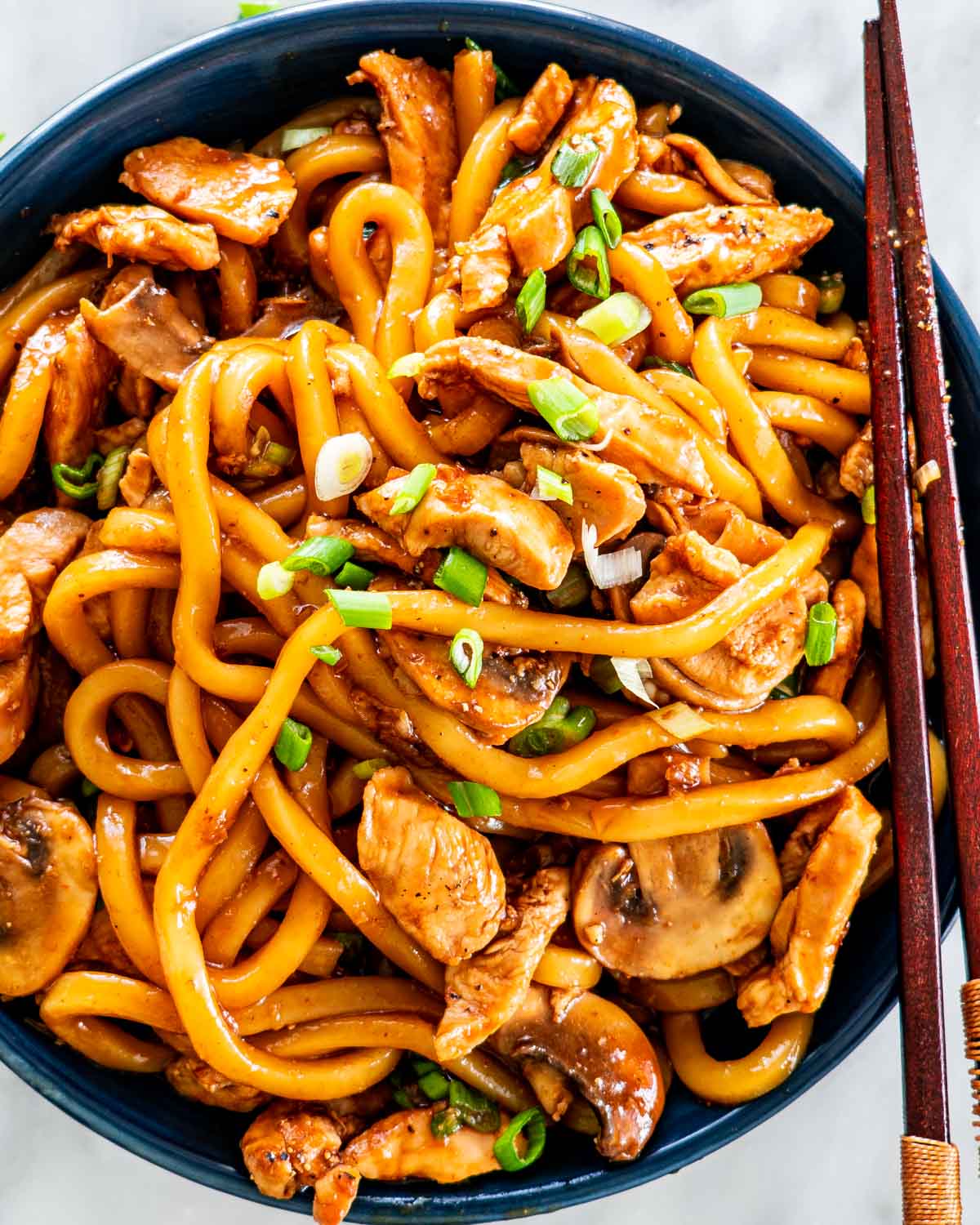 overhead shot of hoisin chicken udon noodles in a blue bowl garnished with green onions and a pair of chopsticks on the plate