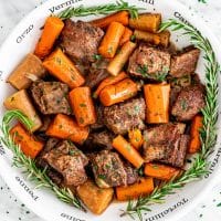 a bowl full of slow cooker short ribs and carrots surrounded by sprigs of rosemary