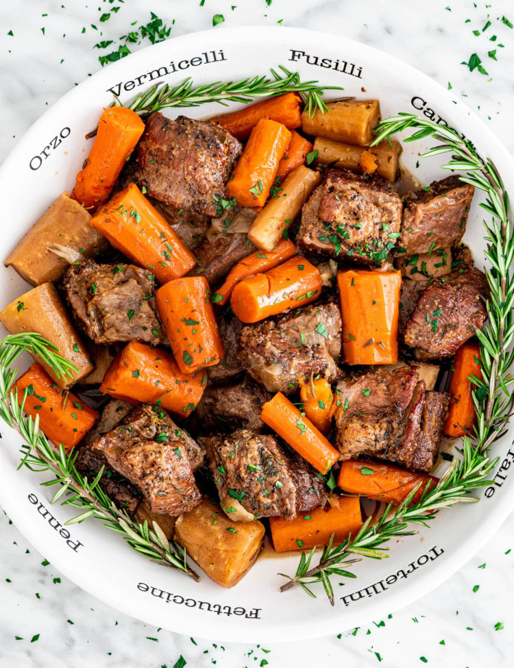 a bowl full of slow cooker short ribs and carrots surrounded by sprigs of rosemary