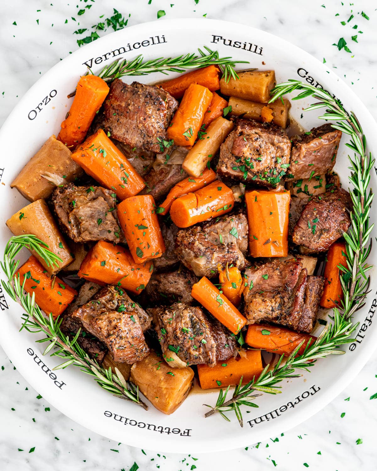 overhead shot of short ribs with carrots and parsnips in a large white bowl garnished with rosemary
